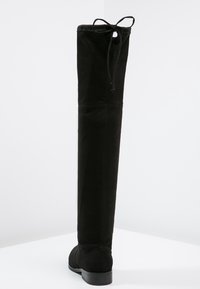 Bota negra por encima de la rodilla hecha de ante con una textura suave. Presenta una punta redondeada, un tacón bajo y un detalle de lazo en la parte superior.