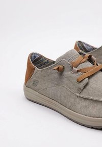 Zapatillas de lona gris claro con acentos de cuero marrón, presentan una punta redonda, diseño con cordones y forro de patrón. Estética simple y moderna.