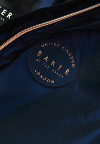Innenansicht einer Marinejacke mit einem kreisförmigen schwarzen Etikett mit metallisch goldener Schrift: "BAKER by Ted Baker, London, Vereinigtes Königreich." Reißverschlussdetail angezeigt.