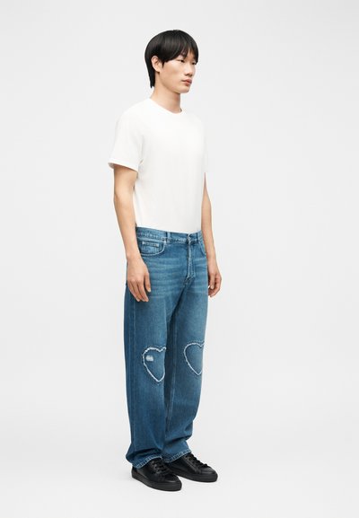 Άνδρας μοντέλο φοράει μια απλή λευκή κοντομάνικη μπλούζα, φαρδιά μπλε τζιν με μπαλώματα σε σχήμα καρδιάς στα γόνατα και μαύρα αθλητικά παπούτσια, στέκεται μπροστά σε αφαιρετικό φόντο.