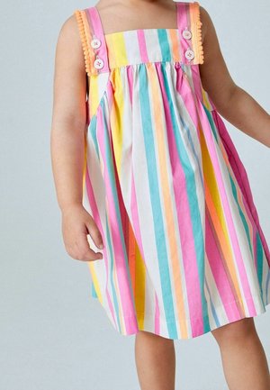 Bambina che indossa un vestito senza maniche con strisce verticali colorate e bordo di pompon sulle spalline, in piedi davanti a uno sfondo semplice.