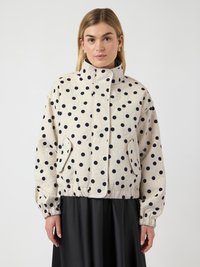 Ξανθιά γυναίκα που φοράει μπεζ μπουφάν καπιτονέ με μαύρες βούλες, στέκεται μπροστά σε λευκό φόντο.