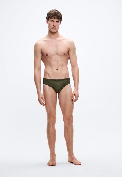 Πράσινα μαγιό σλιπ από λείο υλικό, με κλασική κοπή και μινιμαλιστικό σχέδιο. Φορεμένα από έναν άνδρα μοντέλο με ουδέτερη στάση.