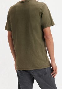 Oliwkowy t-shirt z krótkim rękawem, z okrągłym dekoltem i bezszwowymi ramionami; gładka faktura i luźny krój, zestawiony z szarymi spodniami.