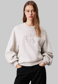 Sudadera gris claro hecha de un tejido suave, con cuello redondo, puños acanalados y un logotipo en relieve en la parte delantera. Combinada con pantalones negros.