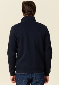 Felpa navy con colletto alto, polsini e orlo a coste. Presenta cuciture a contrasto lungo le spalle e i lati. Abbinata a jeans blu.