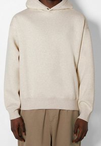 Beige hoodie i ett mjukt, texturerat tyg med dragsko i huvan, ribbade ärmslut och bred nederkant, kombinerad med ljusbeiga byxor.