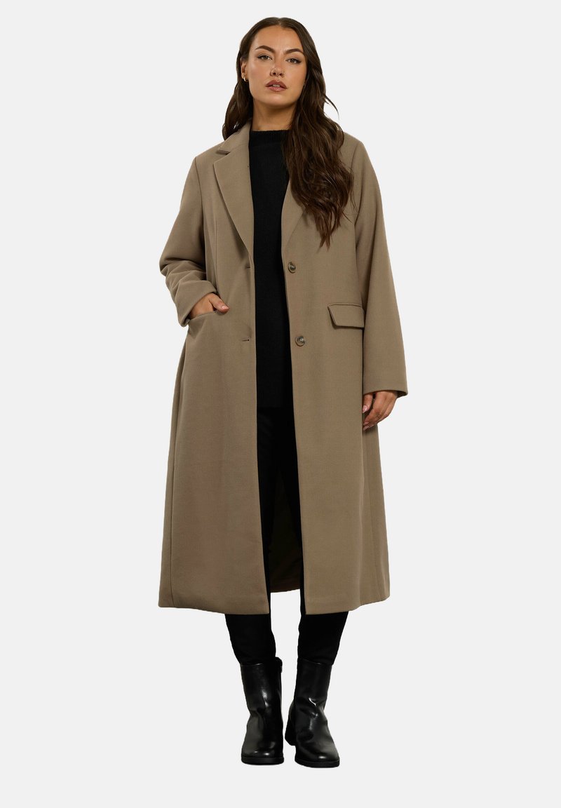 Donna con capelli lunghi che indossa un lungo cappotto beige sopra un outfit nero e stivali neri, in piedi con una mano nella tasca del cappotto.