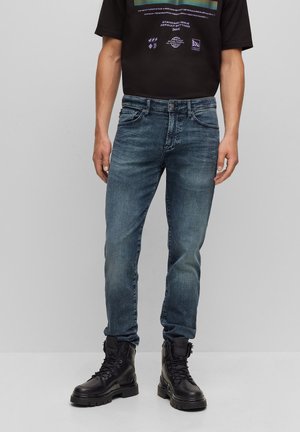 Homme portant un jean slim bleu délavé, des bottes de combat noires et un t-shirt noir à motif graphique, debout devant un fond gris uni.