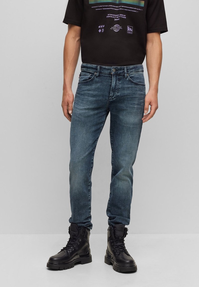 Homme portant un jean slim bleu délavé, des bottes de combat noires et un t-shirt noir à motif graphique, debout devant un fond gris uni.
