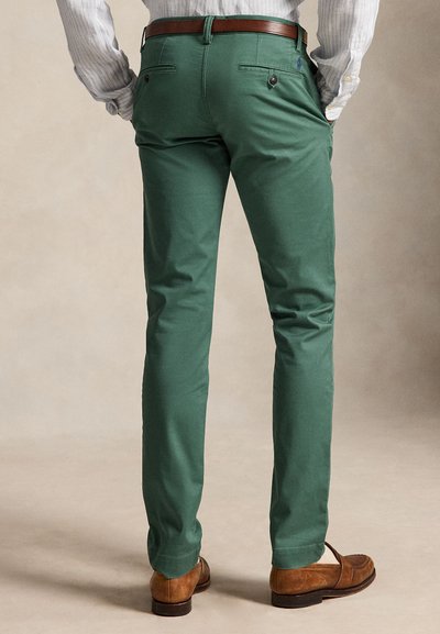 Mand iført grønne slim fit bukser, brunt læderbælte, brune loafers og en stribet langærmet skjorte, stående med hænderne i lommerne.