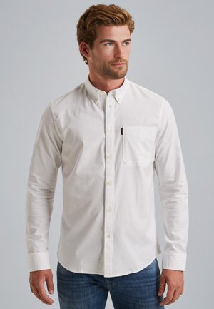 Camisa blanca de manga larga con botones y cuello, un bolsillo en el pecho y botones en el frente, combinada con jeans azules. Textura de tela suave.