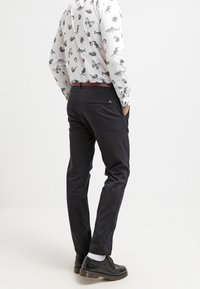 Man standing with hands in pockets wearing black pants, patterned white shirt with animal prints, brown belt, white socks, and black shoes.