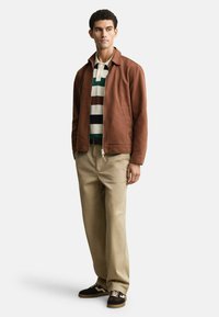 Bruine katoenen jas met een kraag, gestreepte polo in groen, wit en bruin, beige chinos en bruin sneakers met witte accenten.