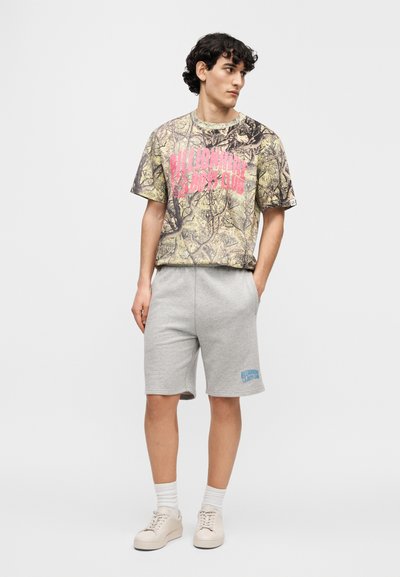 Κοντομάνικο πουκάμισο με καμουφλάζ σχέδιο και ροζ λογότυπο, συνδυασμένο με γκρι σορτς που έχουν μπλε λογότυπο. Το σύνολο ολοκληρώνουν casual παπούτσια.