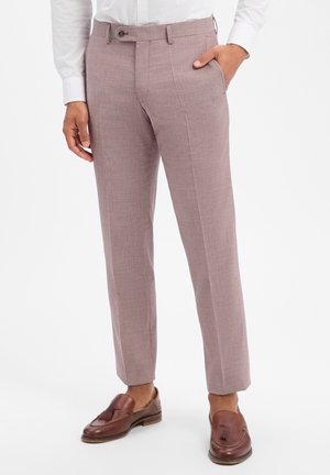 Mann trägt schlanke hellviolette Anzughose, weißes Hemd und braune Lederloafers, steht mit einer Hand in der Tasche.