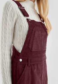 Burgunderfarbene Corduroy-Latzhose mit verstellbaren Trägern, ausgestattet mit einer Brusttasche und silberfarbenen Beschlägen, getragen über einem weißen Zopfmuster-Pullover.