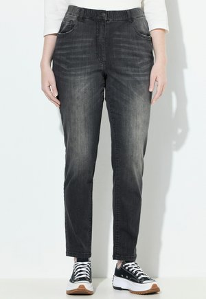 Person, der ausgebleichte schwarze gerade geschnittene Jeans und schwarze und weiße Plateausneaker trägt, steht vor einem weißen Hintergrund.