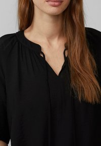 Blouse noire avec un décolleté en V et des liens décoratifs à l'avant. Le matériau semble léger, avec une texture douce et un léger éclat.