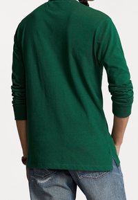 Person klädd i en mörkgrön långärmad tröja och blå slitna jeans, sedd bakifrån med händerna i fickorna.