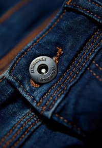 Tummansinisestä farkkukankaasta, jossa on kontrastina oranssia ompelulankaa, ja pyöreä musta metallinen nappi, jossa on kohokuvioitu "LINDENBERGH". Lähikuvaties.