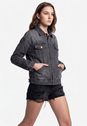 Young woman with long hair wearing a dark washed denim jacket and black ripped shorts, standing with hands in jacket pockets.