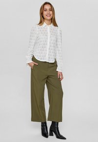 Witte, gebloemde blouse met lange mouwen en een kraag, gecombineerd met olijfgroene wijde broek en zwarte enkellaarsjes.
