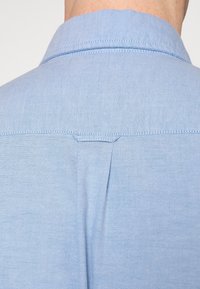 Vaaleansininen paita, jossa on klassinen kaulus, hillitty tekstuuri ja pystysuora sauma yksityiskohtana takana. Pehmeä, hengittävä kangas.