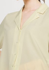 Blouse jaune clair à manches courtes avec un col en V, une patte de boutonnage à l'avant, et un tissu doux texturé. Dotée d'un col et d'un léger éclat.