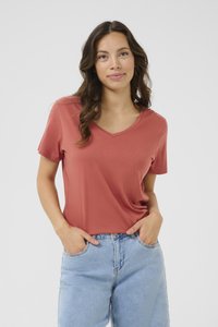 Tričko s výstrihom do V a krátkym rukávom vo farbe koral, vyrobené z mäkkej tkaniny. Kombinované s džínami svetlomodrej farby s vysokým pásom. Model stojí s rukami v vreckách.