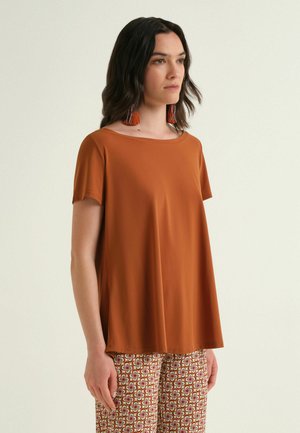 Donna con capelli lunghi e scuri indossa una maglietta a maniche corte color ruggine, pantaloni con motivo geometrico e orecchini con nappine arancioni, in piedi davanti a uno sfondo uniforme.