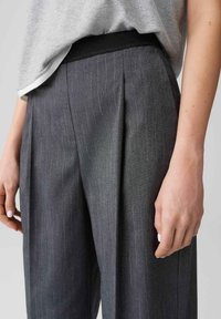 Grijs pantalon met pinstripe, een geplooide voorkant, elastische tailleband en een zachte textuur, weergegeven vanaf de zijkant met een hand op de stof.