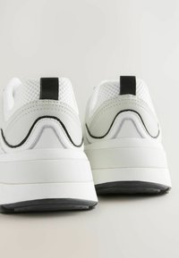 Baskets de sport blanches avec une tige en maille, semelle épaisse blanche, accents noirs et détails texturés sur le talon et la semelle extérieure.