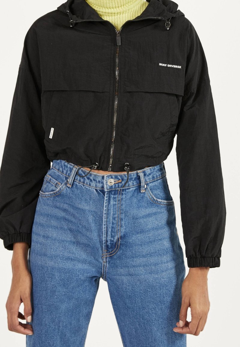 Black cropped jacket with a hood, front zipper, and two upper pockets. Worn over a light green turtleneck and blue high-waisted jeans.