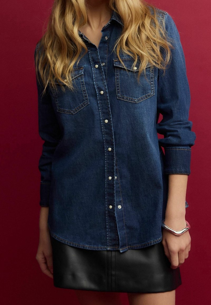 Femme portant une chemise en denim bleu foncé à boutons-pression, une jupe en cuir noire et un bracelet argenté au poignet droit, sur fond rouge.