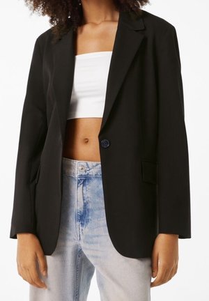 Femme portant un blazer noir oversize sur un top tube blanc et un jean bleu clair taille haute, debout avec les bras détendus.