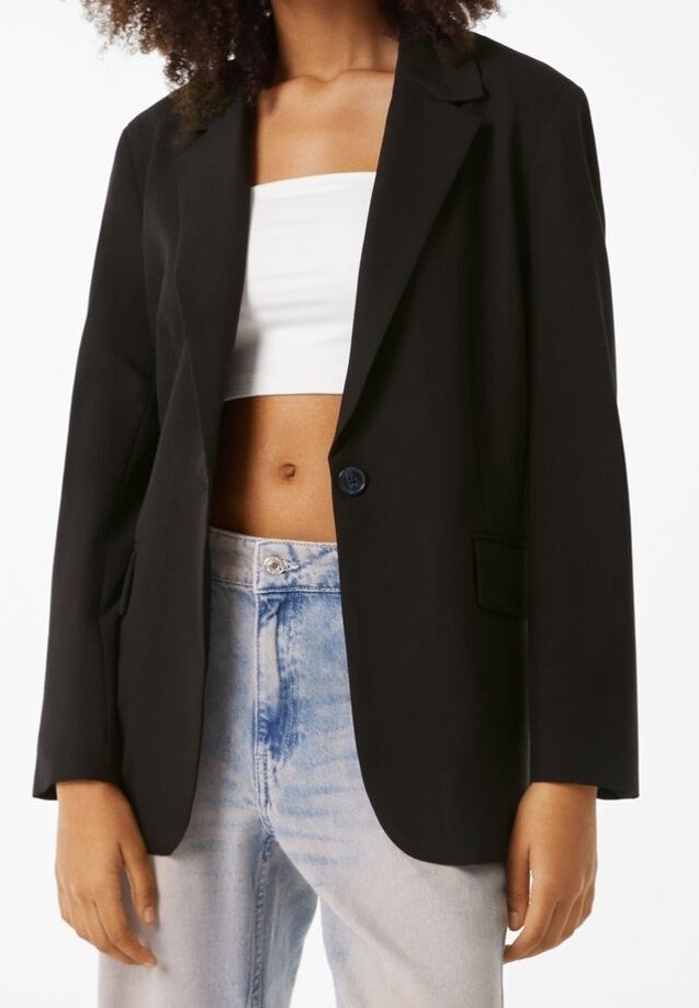 Femme portant un blazer noir oversize sur un top tube blanc et un jean bleu clair taille haute, debout avec les bras détendus.