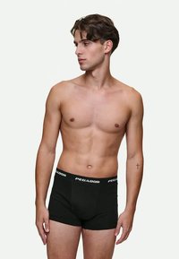 Hombre joven con pelo corto castaño, vistiendo un bóxer negro Pegador, de pie sin camiseta y mirando hacia su izquierda contra un fondo liso.