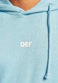Lys blå hoodie med en frontlomme, snorehætte og et hvidt trykt "DEF" logo i midten. Blød tekstur, afslappet design.