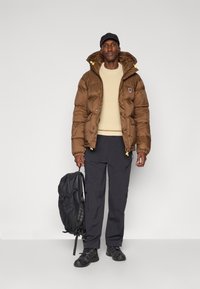 Brown puffer jacket, textured fabric, with yellow accents, worn over a cream knit sweater; paired with black pants and a black backpack.