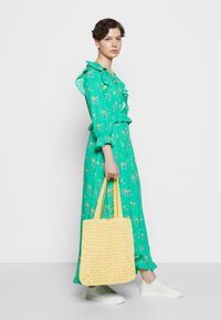 Vrouw in een groene bloemrijke maxi-jurk die een grote gele gevlochten tas vasthoudt, gekleed in witte sneakers, staande tegen een effen witte achtergrond.