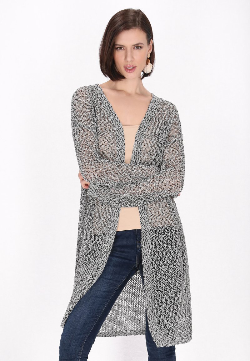 Leichtes, offen gestricktes Cardigan in Grau und Schwarz. Mit einem tiefen V-Ausschnitt und langen Ärmeln. Wird über einem beigen Oberteil mit engen dunklen Jeans getragen.