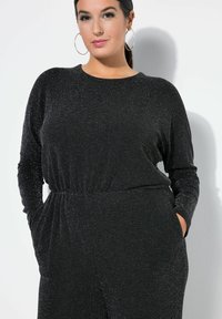 Mono negro texturizado con mangas largas y cintura elástica. Presenta hilos metálicos y bolsillos laterales. Cuello redondo.