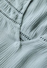Lichtblauwe stof met een gekreukt textuur, voorzien van horizontale rijen decoratieve stiksel en kleine uitsparingen langs de naad.