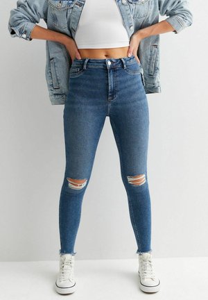 Osoba ubrana w wysokie, podarte niebieskie skinny jeansy, białą crop top, dżinsową kurtkę i białe trampki, stoi z rękami na biodrach.