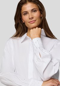 White button-up shirt with long, voluminous sleeves. Cuffs adorned with small, clear decorative studs. Smooth, lightweight fabric.