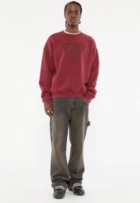 Bordeaux sweatshirt met groot zwart driehoeklogo, ontspannen pasvorm, geribde manchetten. Gecombineerd met losse, donkergrijze jeans en zwarte schoenen.