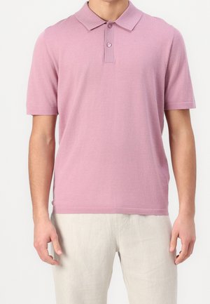 Homme portant un polo rose clair à manches courtes avec un col et deux boutons, associé à un pantalon beige clair.