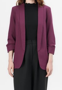 Blazer violet au design élégant, avec des revers structurés et des manches retroussées, porté sur un haut noir côtelé. Associé à un pantalon noir.