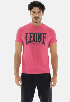 Homme aux cheveux foncés portant un t-shirt rose ajusté avec "LEONE 1947" en grandes lettres noires et un pantalon noir, debout devant un fond blanc.